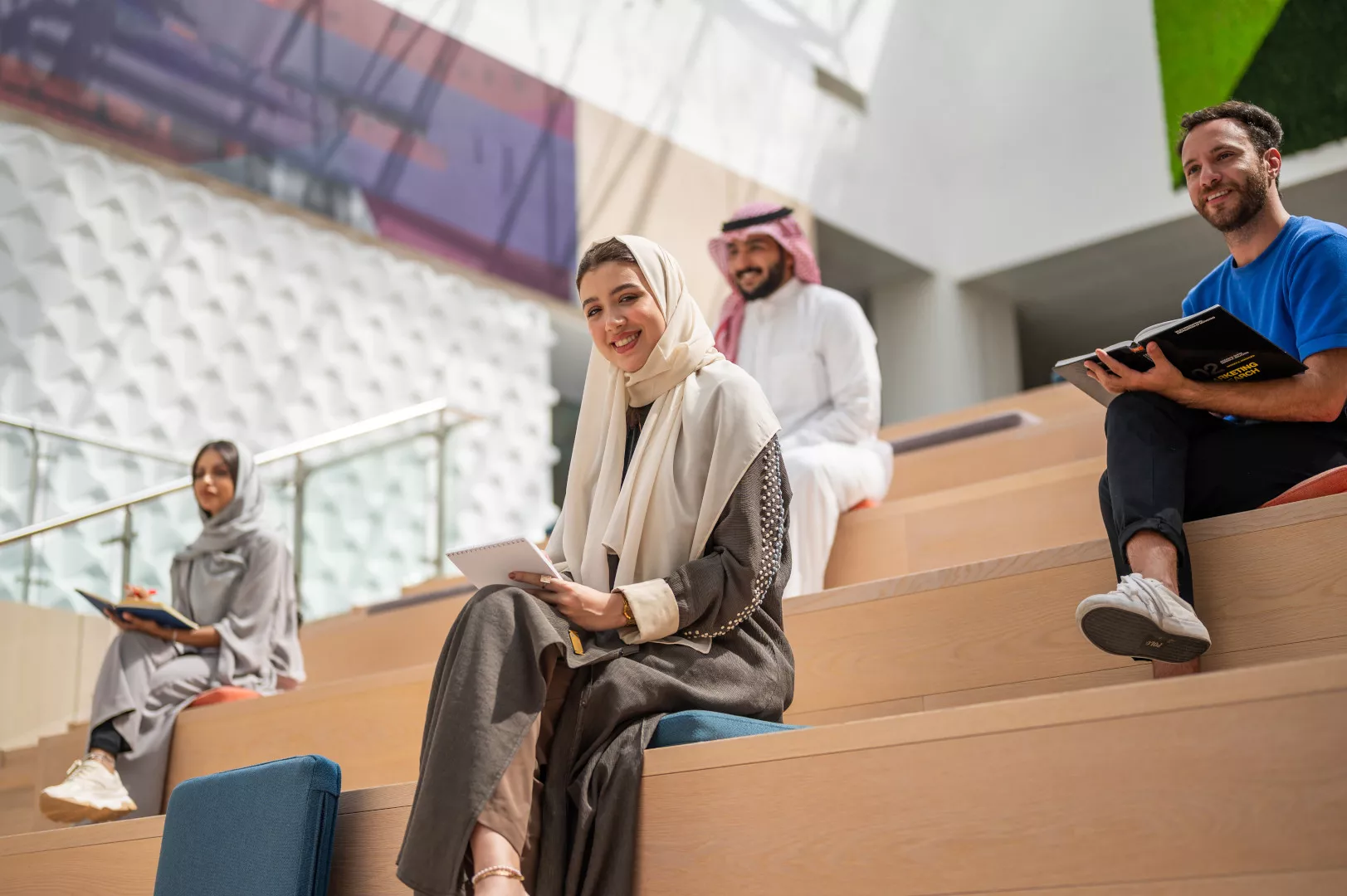 A group of diverse young individuals are seated on modern wooden steps in a bright, contemporary setting. In the foreground, a young woman in a beige hijab and abaya smiles, holding a notebook. Behind her, a man in traditional Middle Eastern attire and another woman in a grey hijab are visible. To the right, a man in a blue t-shirt holds an open book, smiling.